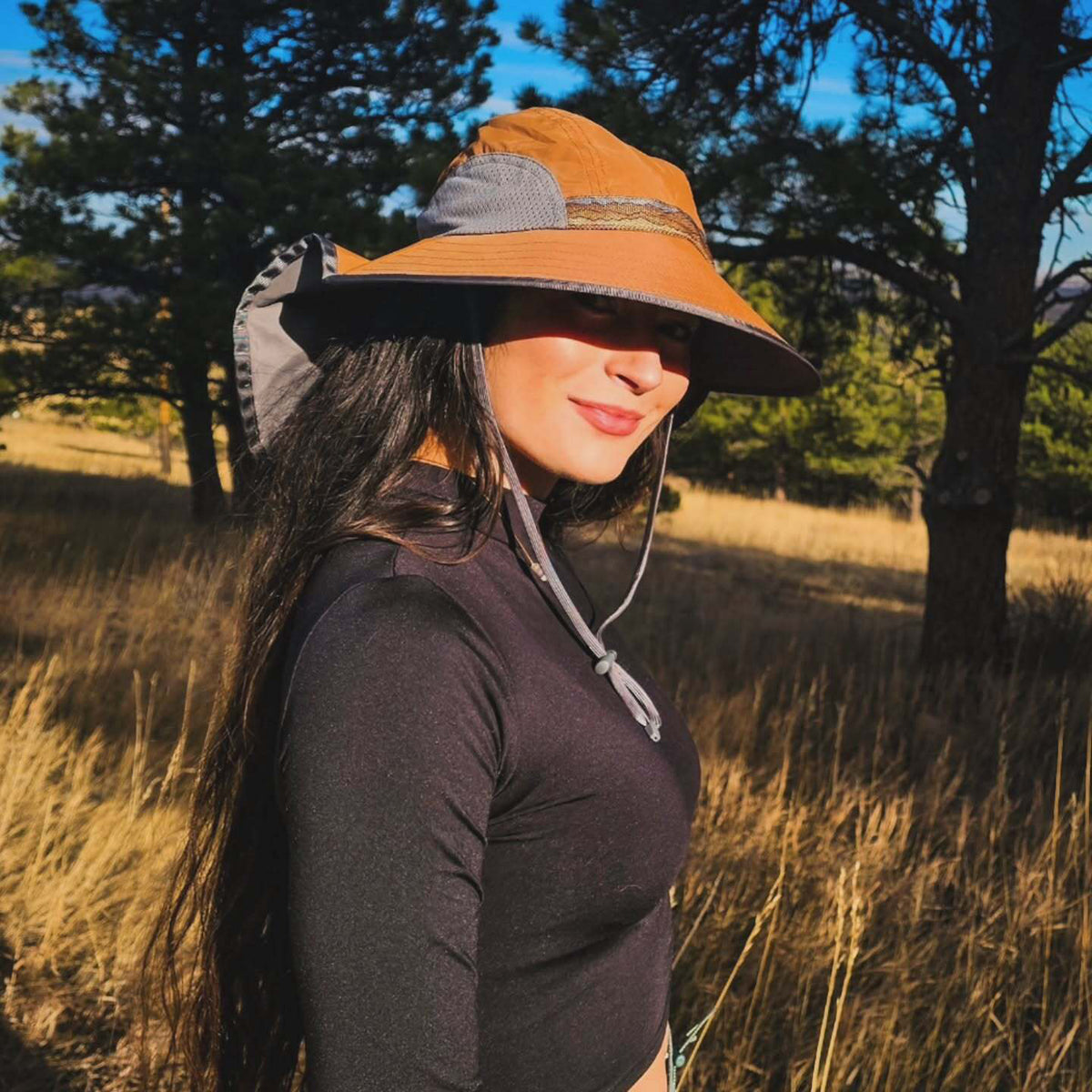 A woman wearing a wide-brimmed brown and gray sun hat with a chin strap, standing in a grassy outdoor field with trees in the background.