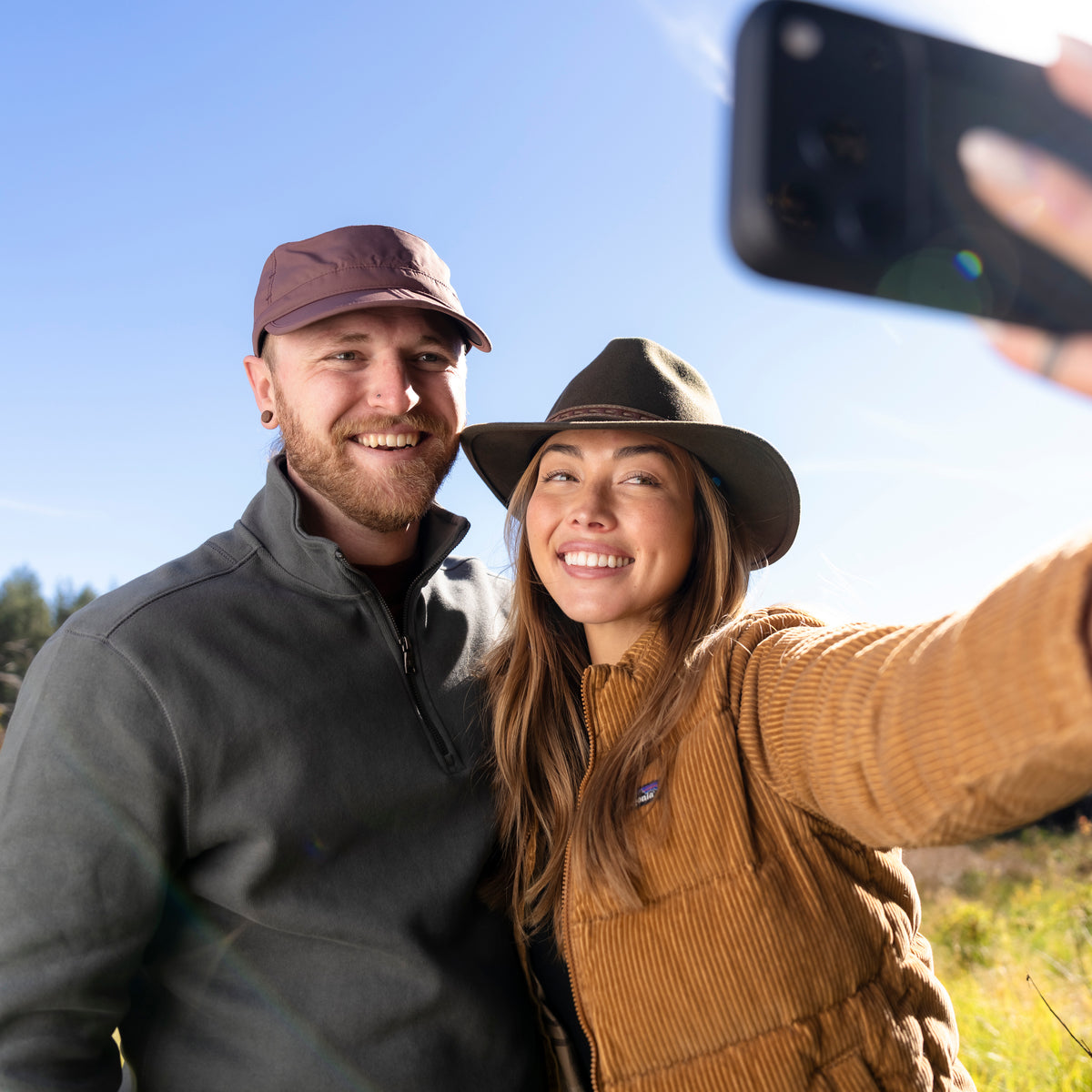 A man and a woman taking a sunny outdoor selfie. The man wears a brown cap and a dark gray fleece pullover, while the woman wears a brown wide-brim hat and a tan corduroy vest over a brown top.