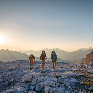 A photo of three people hiking towards the camera on some rocky terrain with mountains and a sunrise in the background.