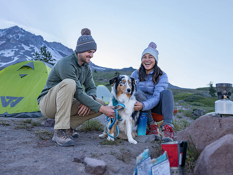 A man and a woman camping in the fall, wearing beanies crouched down petting a dog
