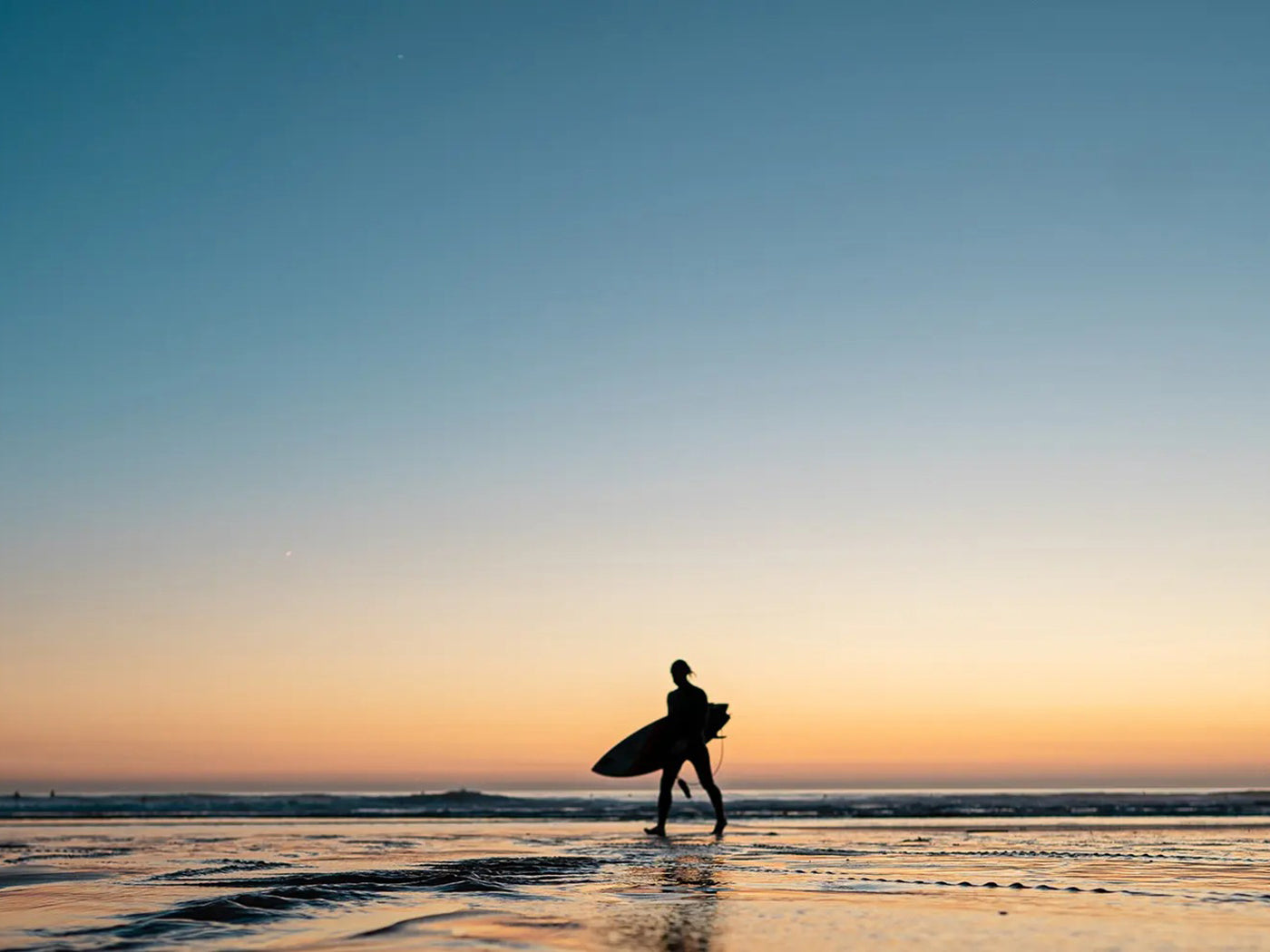 Person in the distance holding a surfboard on the beach at sunrise