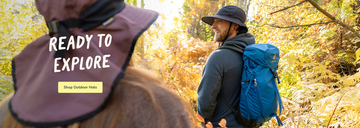 A man wearing a dark wide-brim hat and a blue hiking backpack walks through a sunlit autumn forest, while another person in the foreground wears a maroon sun hat with the text 'READY TO EXPLORE' and a 'Shop Outdoor Hats' button.