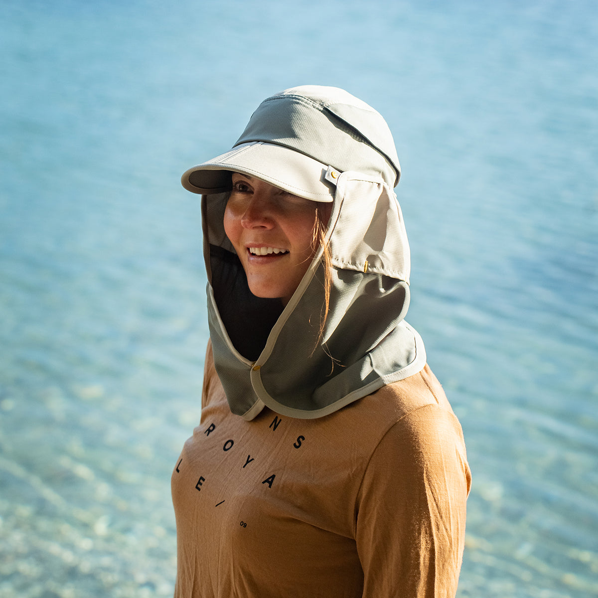 A woman wearing a beige sun hat with an attached neck flap and chin shield, standing outdoors with a blue body of water in the background.