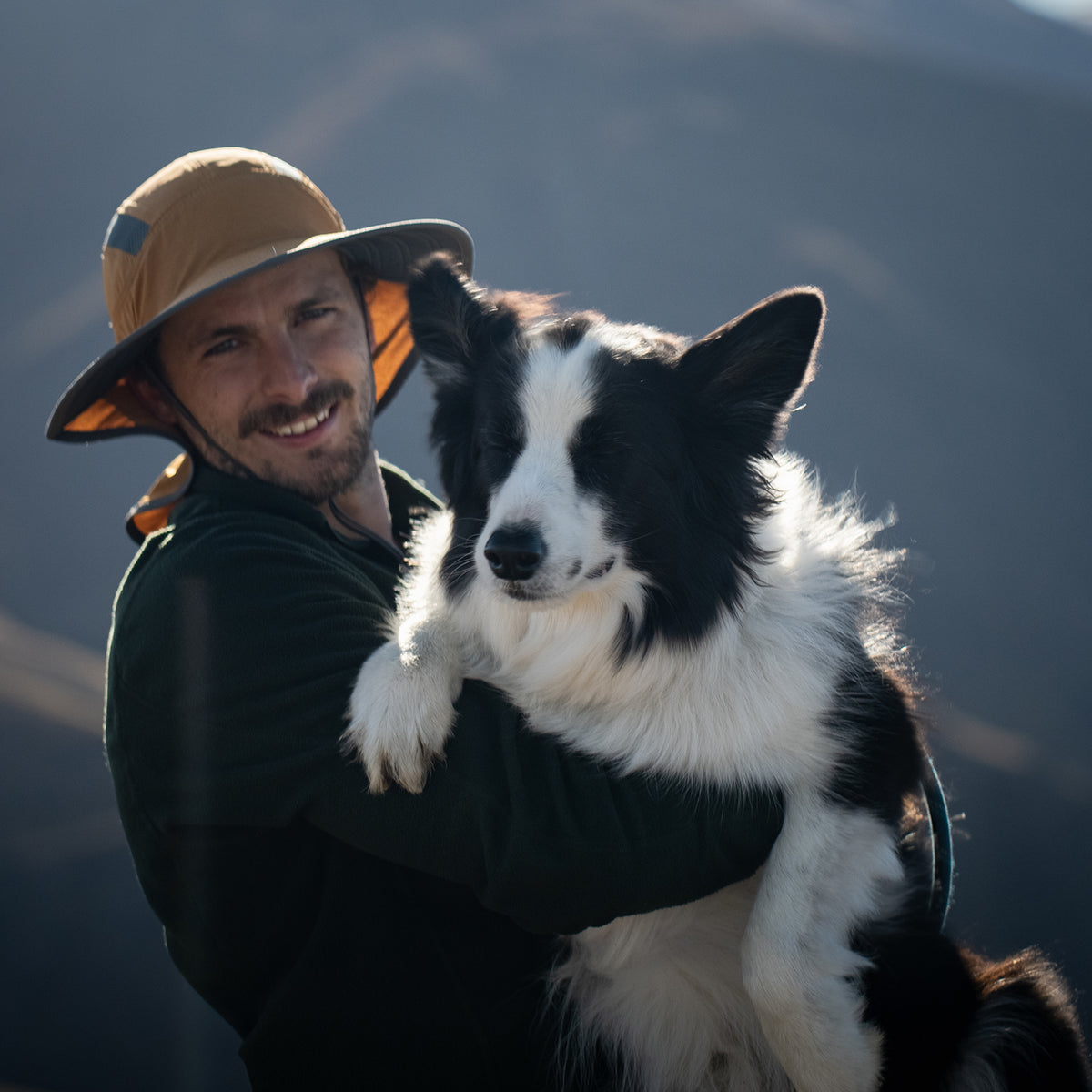 A smiling man wearing a tan wide-brim hat and dark green shirt holds a black and white Border Collie outdoors against a blurred mountain background.