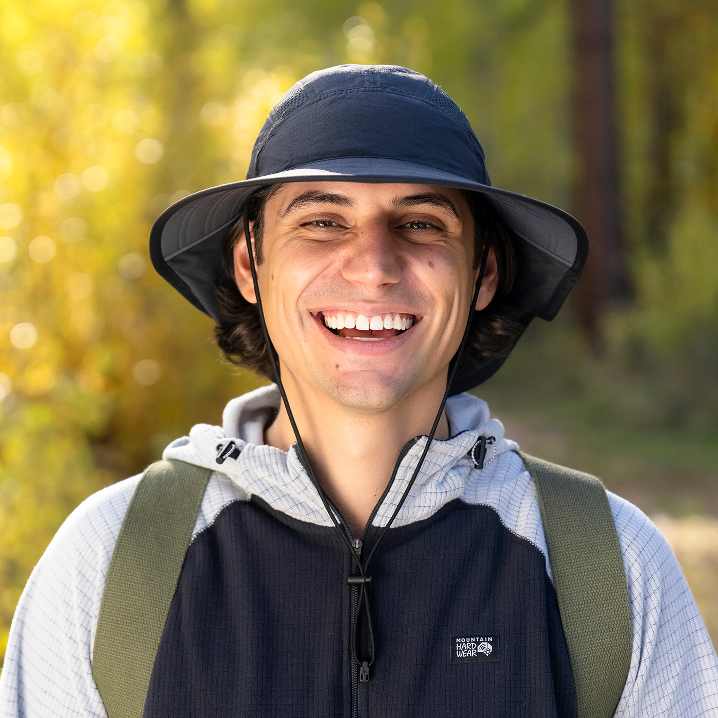 A smiling man outdoors wearing a dark navy wide-brim bucket hat, a navy and white zip-front vest over a white hoodie, with green backpack straps visible, against a blurred forest background.